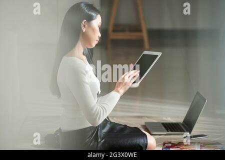 Jeune femme asiatique sérieuse occupée avec des cheveux longs noirs assis sur le sol avec des gadgets et utilisant une tablette tout en travaillant avec le stockage dans le cloud Banque D'Images