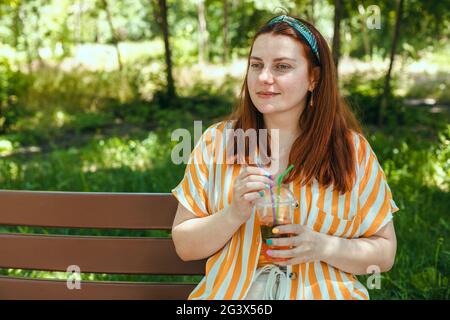 Été gaie femme avec un cocktail froid assis sur un banc en bois dans le parc de la ville, appréciant le reste. Concept d'alimentation saine Banque D'Images