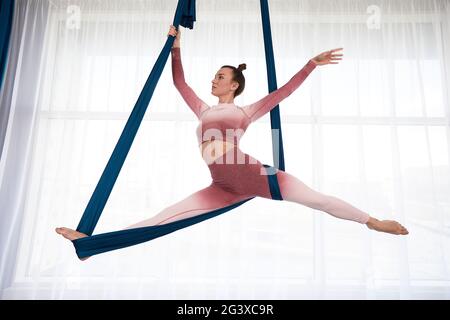 Une fille est engagée dans le yoga aérien faisant des exercices d'étirement de ficelle dans un hamac contre une grande fenêtre. Banque D'Images