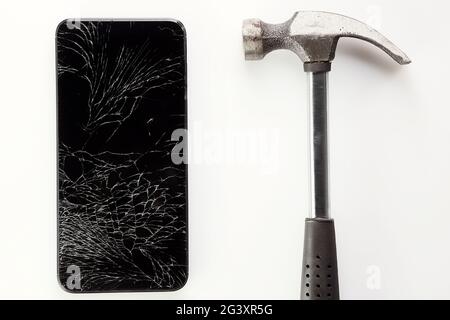 Un smartphone noir cassé avec un écran fissuré et un marteau en métal se trouve sur un fond blanc. Banque D'Images