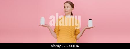 Brunette fille tenant des bouteilles médicales de médicaments, les vitamines des deux côtés ouvrir la paume faisant le choix difficile contre le concept de traitement de fond rose Banque D'Images