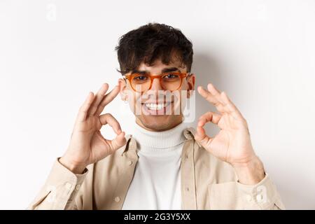 Gros plan d'un beau gars dans de nouvelles lunettes montrant des signes corrects et souriant heureux, louant de bonnes lunettes, debout sur le backgro blanc Banque D'Images