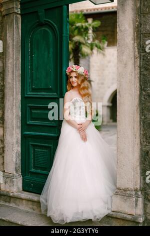 Une mariée avec une couronne de roses se tient à la porte verte dans la cour intérieure confortable de la vieille ville De Kotor Banque D'Images