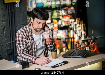 Propriétaire d'une petite entreprise au téléphone et à l'ordinateur dans le magasin. Un travailleur de la boutique de sport fait un appel avec un téléphone cellulaire. Vendeur travaillant et parlant Banque D'Images