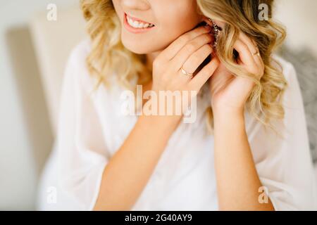 La mariée portant un peignoir blanc met sur une boucle d'oreille et sourit tout en préparant le mariage Banque D'Images