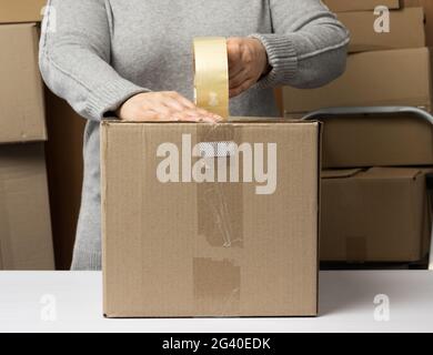 Femme dans un chandail gris tient un rouleau de ruban adhésif et paquet des boîtes en carton brun sur une table blanche Banque D'Images