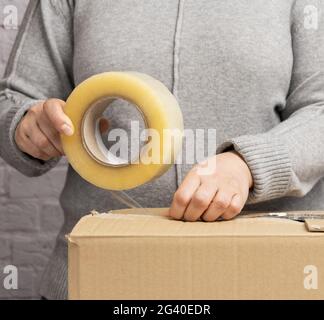 Femme dans un chandail gris tient un rouleau de ruban adhésif et paquet des boîtes en carton brun Banque D'Images