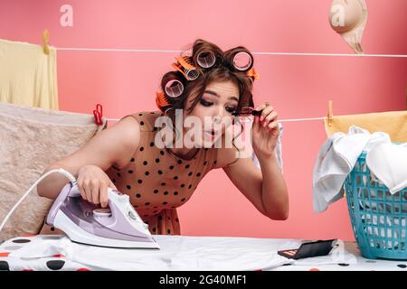 Femme, femme au foyer repassant des vêtements sur une planche à découper et faisant du maquillage en même temps. Corde avec des vêtements sur fond rose. Banque D'Images