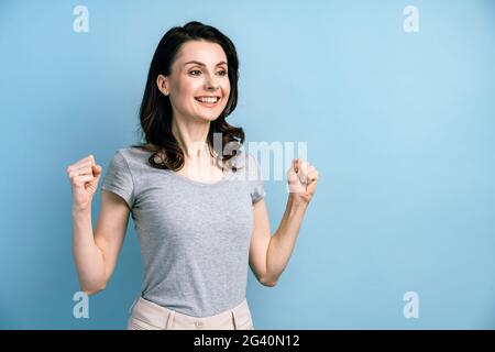 Belle dame attrayante avec des poings élevés, se réjouit sincèrement de sa victoire. Fille isolée sur fond bleu avec espace de copie pour le texte. Banque D'Images