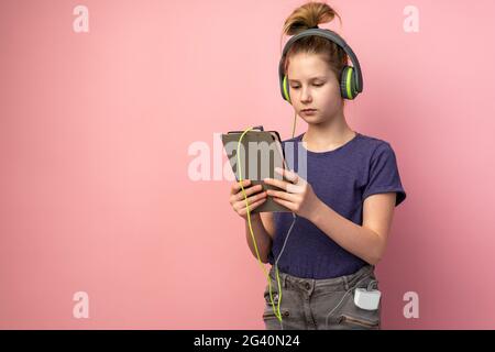 Une adolescente sérieuse dans un casque et une tablette dans les mains sur fond rose. Un enfant mignon apprend ou regarde quelque chose sur l'appareil. Banque D'Images