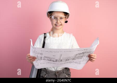 Jolie petite fille dans un casque de construction sur fond rose. Adolescent tenant un dessin dans ses mains. Banque D'Images