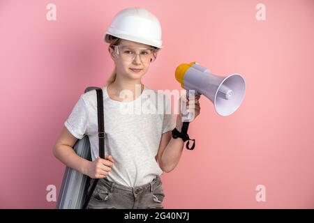 Concept de conception. Jolie petite fille dans un casque de construction et des lunettes et un haut-parleur dans ses mains sur fond rose. Banque D'Images