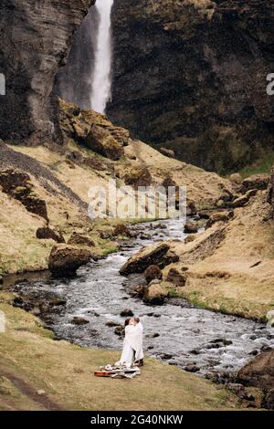 Destination mariage Islande, près de la cascade de Kvernufoss. Un couple de mariage se tient sous un plaid près d'une rivière de montagne. Le marié h Banque D'Images