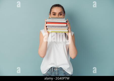 Fille préoccupée étudiant tenant des cahiers et des livres. Isolée sur fond bleu, l'élève couvre la moitié de son visage avec ses notes. Banque D'Images