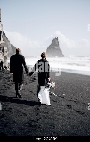 Destination mariage Islande. Un couple de mariage marche le long de la plage de sable noir de Vik, près de la roche de basalte, sous forme de pil Banque D'Images