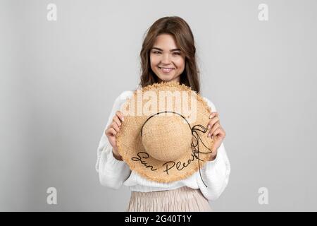 Souriante, belle fille tenant un chapeau de paille dans ses mains. Mignonne, jeune femme isolée sur fond gris, souriante, belle fille tenant un chapeau de paille je Banque D'Images