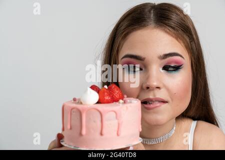 Vue rapprochée. Une jeune fille charmante regarde le gâteau, elle veut vraiment le manger. Jolie fille avec un gâteau sur fond blanc, vue rapprochée. Un vous Banque D'Images