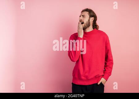 Jeune homme portant un t-shirt décontracté debout sur fond rose isolé. Le bâillement lassant couvre tiredly sa bouche avec sa main. Anxiété et somnolence. Banque D'Images