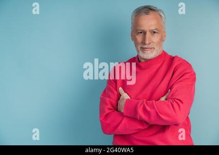 Un homme sérieux, âgé, a traversé les bras sur sa poitrine en posant sur un fond bleu. Homme attrayant isolé sur l'espace de copie Banque D'Images
