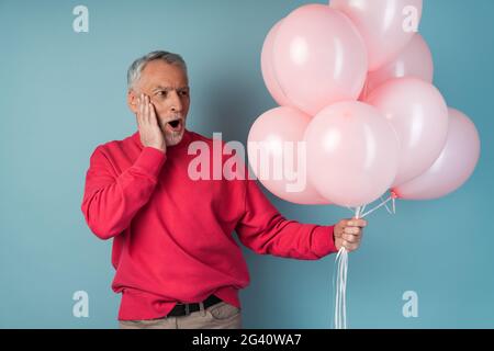 Surpris, l'homme plus âgé tient des ballons roses dans sa main, il les regarde et est surpris. Homme attrayant dans un pull brillant sur un fond bleu, Banque D'Images