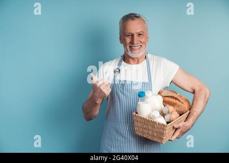 Un homme souriant aux cheveux gris et à la barbe tient un panier de nourriture et pointe son pouce à l'espace de copie. Grand-père mignon posant sur un fond bleu., S. Banque D'Images