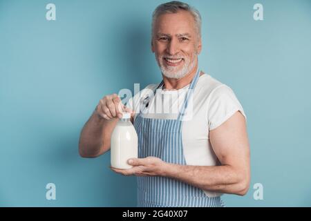 Homme souriant tenant une bouteille de lait. Homme beau, âgé dans un tablier tenant une bouteille de lait sur un fond bleu. Banque D'Images