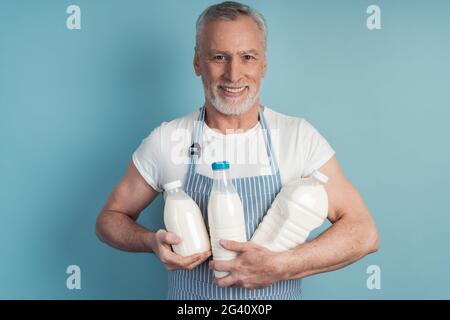 Homme positif, souriant, senior tenant une bouteille de lait sur un fond bleu isolé. L'homme âgé porte un tablier Banque D'Images