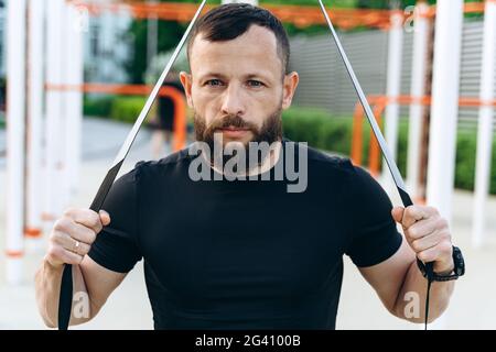 Un homme sérieux avec une barbe est engagé dans l'air libre. L'homme est engagé dans la coupe transversale, effectue continuellement des exercices, regarde la caméra Banque D'Images