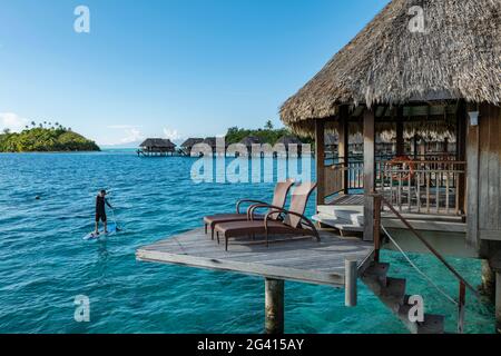 Le poste de paddle-up Man on SUP passe devant un bungalow sur l'eau du Sofitel Bora Bora Private Island Resort à Bora Bora Lagoon, Bora Bora, Leeward Islands, Banque D'Images