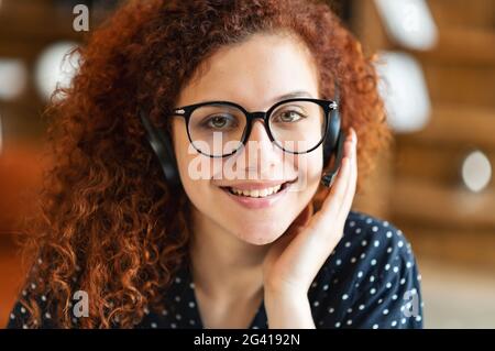 Belle femme à la tête rouge portant un casque sans fil et des lunettes stylées regarde la caméra, prend un appel et souriant, portrait en gros plan d'un représentant de l'assistance responsable en contact, vue webcam Banque D'Images