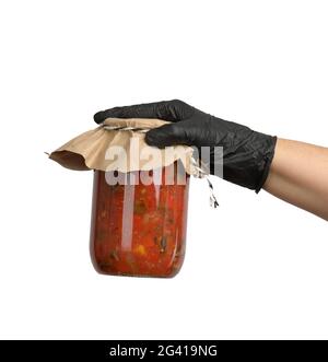 La main femelle dans un gant en latex noir tient un pot en verre avec des tomates en conserve dans la sauce Banque D'Images