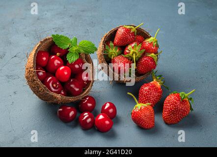 Deux tasses de noix de coco sont remplies de cerises mûres et juteuses et de fraises sur fond bleu foncé. Vue latérale, gros plan. Banque D'Images