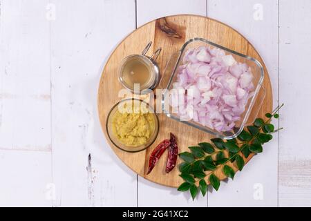 Ingrédients - oignon haché, pâte d'ail au gingembre, feuilles de curry, huile de cuisson et piment sec disposés sur une base en bois avec fond texturé. Banque D'Images