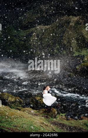 Le marié entoure la mariée dans ses bras près de la cascade, il neige. Destination mariage Islande, près de Kvernufoss Waterfal Banque D'Images