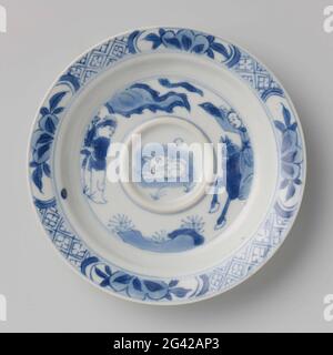 Soucoupe avec le lièvre lunaire et deux figures, une à cheval. Plat de porcelaine, avec une augmentation annulaire sur le plat, peint en bleu sous-glaçure. Sur le plat, le lièvre de lune dans le médaillon surélevé, où autour d'une scène: Un cavalier et un porteur standard dans un paysage; le bord avec la serviette entremêlée avec des cartouilles avec branche de fruits; quatre objets de valeur (feuille d'artemisia, perle, rhombus, rouleau) sur le dos. Marqué sur le dessous avec la marque de quatre caractères de l'empereur Chenghua dans un double cercle. Bleu blanc. Banque D'Images