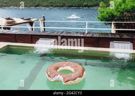 Un cercle gonflable flotte dans une piscine donnant sur la mer, les montagnes et un yacht amarré. Banque D'Images