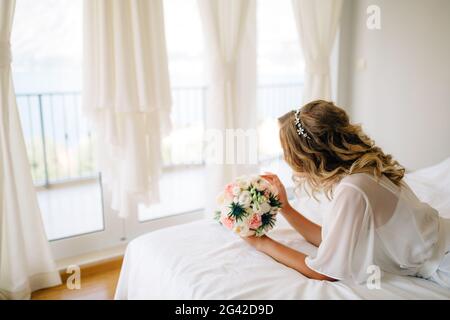 Une mariée dans un peignoir doux avec un bouquet ses mains sur le lit par la fenêtre avec rideaux blancs Banque D'Images