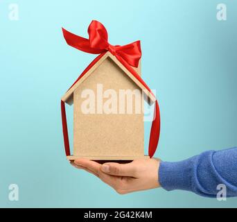 Les mains des femmes tiennent un modèle d'une maison en bois attaché avec un ruban de soie rouge sur un fond bleu Banque D'Images