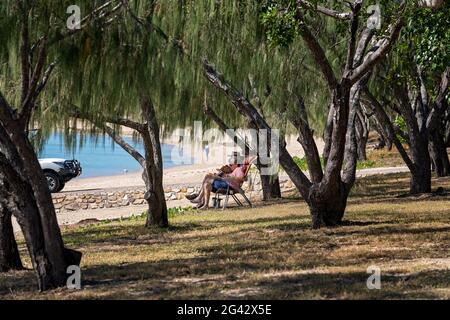 Dingo Beach, Queensland, Australie - juin 2021 : un couple de personnes âgées se détendant et lisant à l'ombre des arbres à côté d'une rampe de chargement de bateau sur la plage Banque D'Images