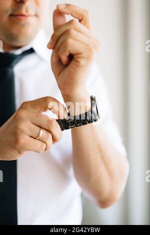 Un homme dans une chemise blanche et cravate met un regardez sur sa main pendant la préparation de la cérémonie de mariage Banque D'Images