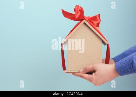 Les mains des femmes tiennent un modèle d'une maison en bois attaché avec un ruban de soie rouge sur un fond bleu Banque D'Images