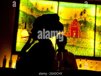 Une femme noire avec un foulard fumant une cigarette , en silhouette contre un panneau de junglescène allumé à un bar. Tourné à Bali. Banque D'Images