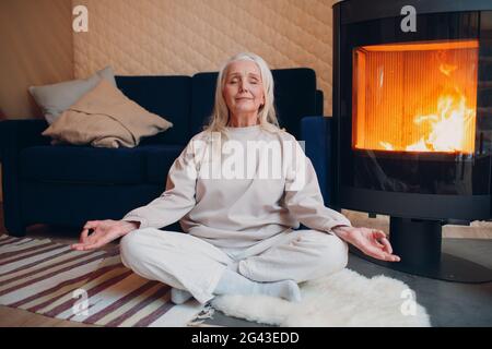 Portrait d'une femme âgée assise dans une position de lotus à l'intérieur avec cheminée. Yoga et méditation zen comme concept. Banque D'Images