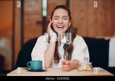 Rire jeune femme heureuse assise à la table à la maison derrière un ordinateur portable et parlant sur appel vidéo. Fille femelle avec portable Banque D'Images