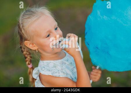 Petite fille blonde mangeant des bonbons en coton bleu dans le parc. Banque D'Images