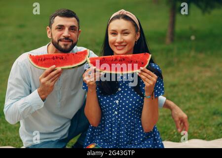 Jeune femme adulte et couple d'hommes pique-nique à l'herbe verte pré dans le parc ayant le plaisir et le sourire avec la pastèque Banque D'Images