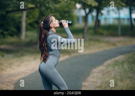 Femme fatiguée jogger en combinaison grise boire de l'eau en bouteille après avoir fait du jogging dans le parc Banque D'Images