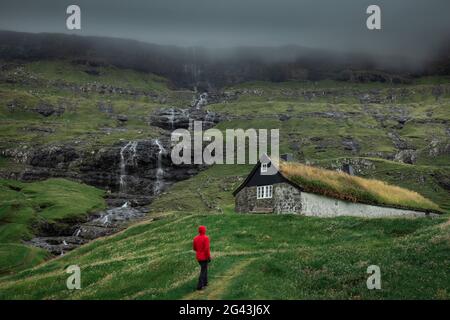 Homme en blouson rouge devant des huttes avec des toits d'herbe dans le village de Saksun sur l'île de Streymoy, îles Féroé Banque D'Images