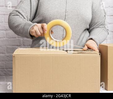 Femme dans un chandail gris tient un rouleau de ruban adhésif et paquet des boîtes en carton brun Banque D'Images