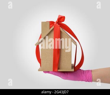 Les mains des femmes tiennent un modèle d'une maison en bois attaché avec un ruban de soie rouge sur un fond blanc Banque D'Images